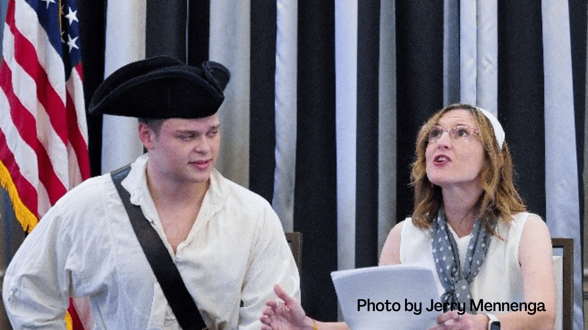 Woman advising man in three-cornered hat in front of an American flag.